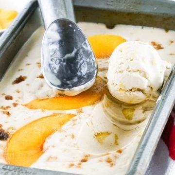 Vanilla ice cream with peach slices in a metal tray, with an ice cream scooper and a bowl of peaches nearby.