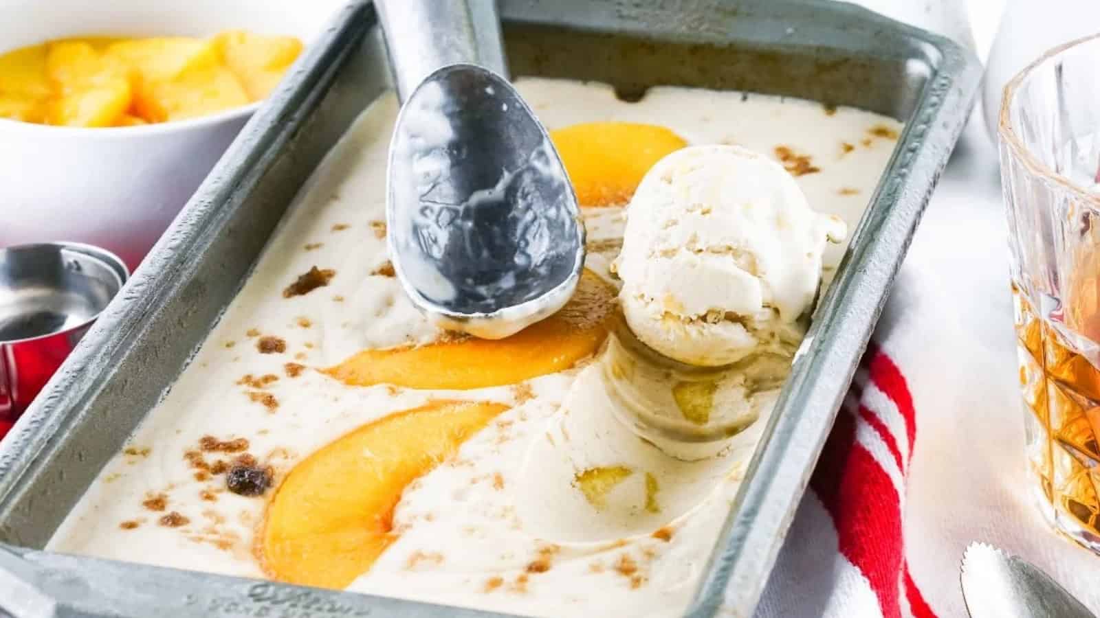 Vanilla ice cream with peach slices in a metal tray, with an ice cream scooper and a bowl of peaches nearby.