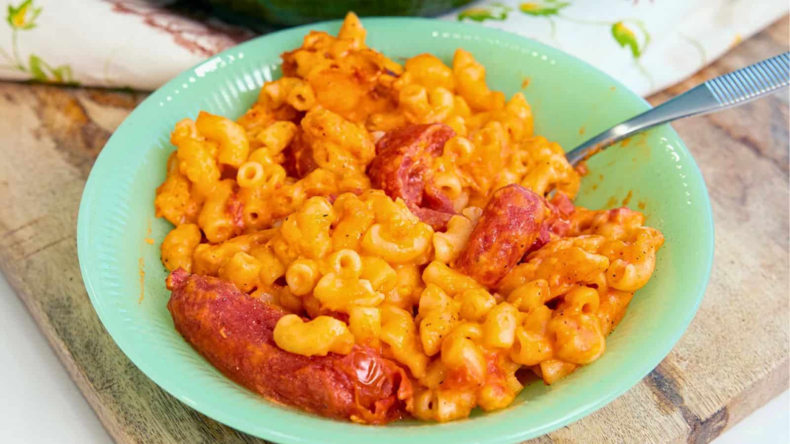 A bowl of macaroni and cheese with slices of sausage, served on a green plate with a fork.