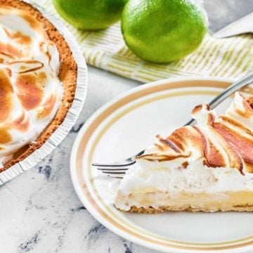A slice of meringue pie on a plate with whole limes and the rest of the pie in the background.