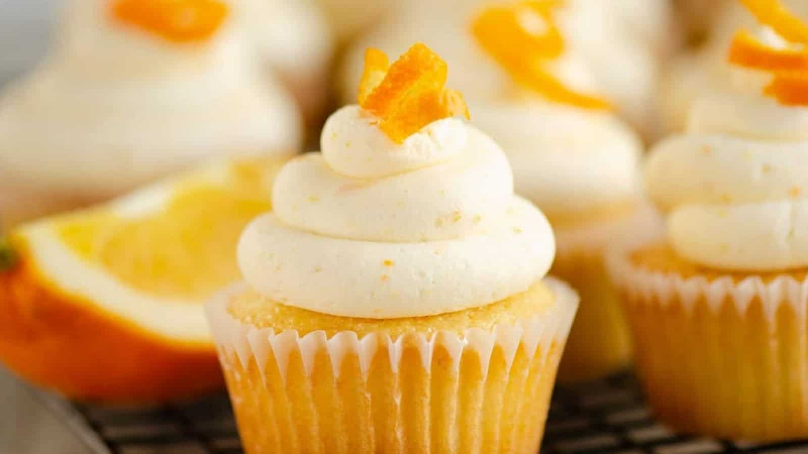 Vanilla cupcakes topped with swirled frosting and orange zest, with an orange slice in the background.