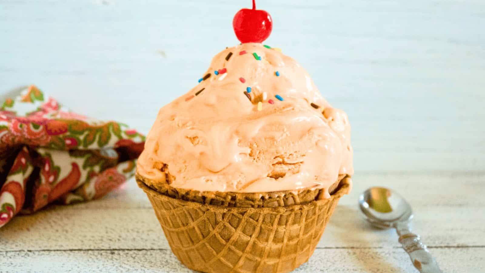 Peach ice cream in a waffle bowl topped with sprinkles and a cherry, next to a spoon and patterned napkin.