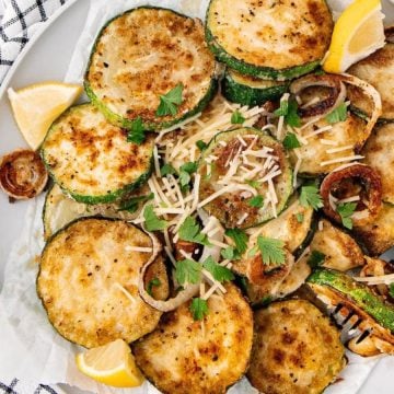 Plate of breaded fried zucchini slices garnished with parsley, lemon wedges, and shredded cheese on a checkered cloth.