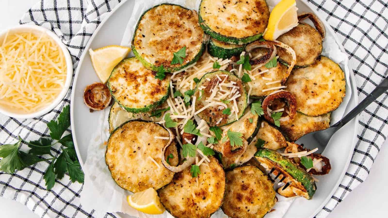 Plate of breaded fried zucchini slices garnished with parsley, lemon wedges, and shredded cheese on a checkered cloth.