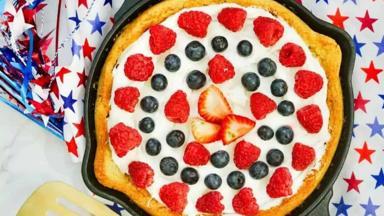 A patriotic fruit pizza topped with raspberries, blueberries, and strawberries in a skillet, with star decorations nearby.
