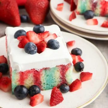 A slice of red, white, and blue poke cake topped with whipped cream, strawberries, and blueberries.