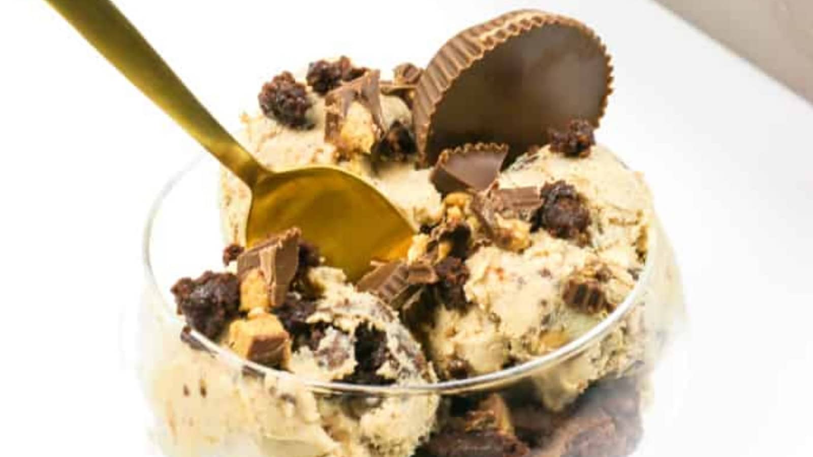 A glass bowl of ice cream topped with peanut butter cups, brownie pieces, and a gold spoon.