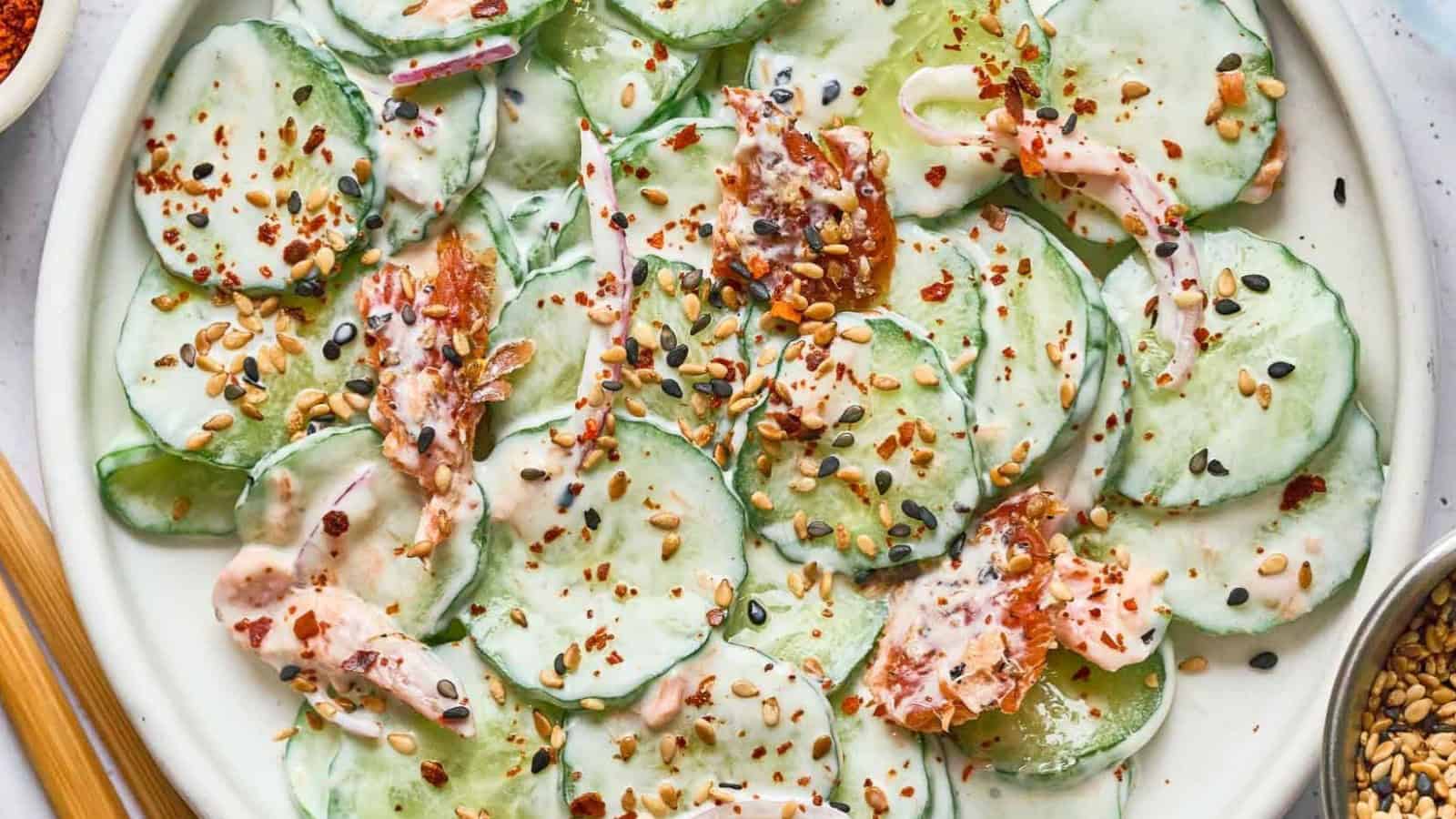 Cucumber salad with creamy dressing, sprinkled with seeds, herbs, and red pepper flakes on a white plate.