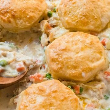 Golden biscuits on creamy chicken pot pie filling with peas and carrots, served in a baking dish with a wooden spoon.