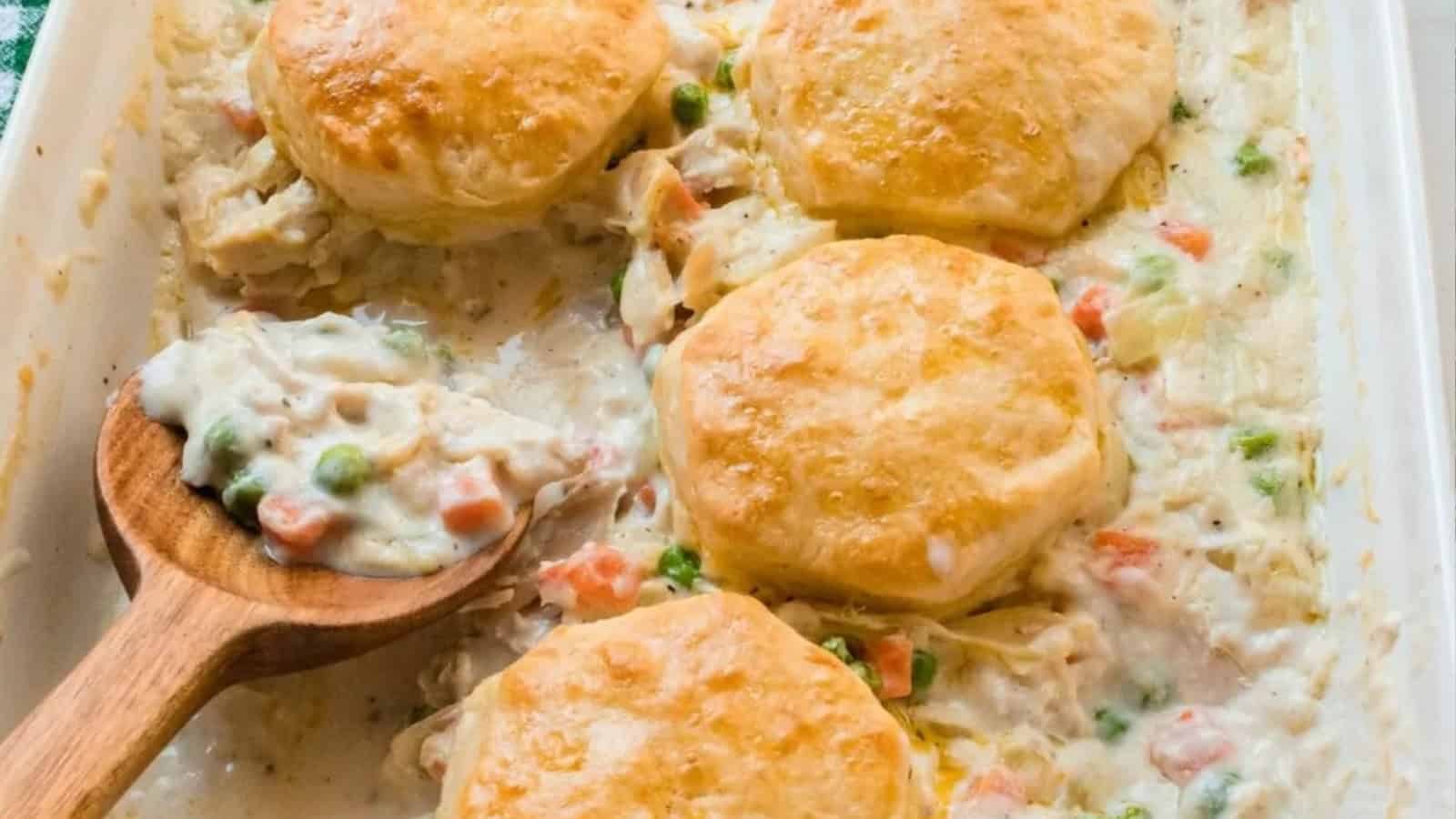 Golden biscuits on creamy chicken pot pie filling with peas and carrots, served in a baking dish with a wooden spoon.
