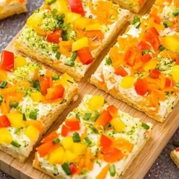 Colorful veggie-topped cracker squares with cream cheese, garnished with chopped chives, on a wooden board.