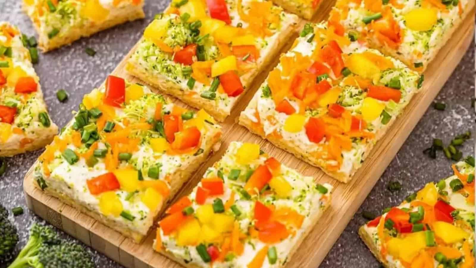 Colorful veggie-topped cracker squares with cream cheese, garnished with chopped chives, on a wooden board.