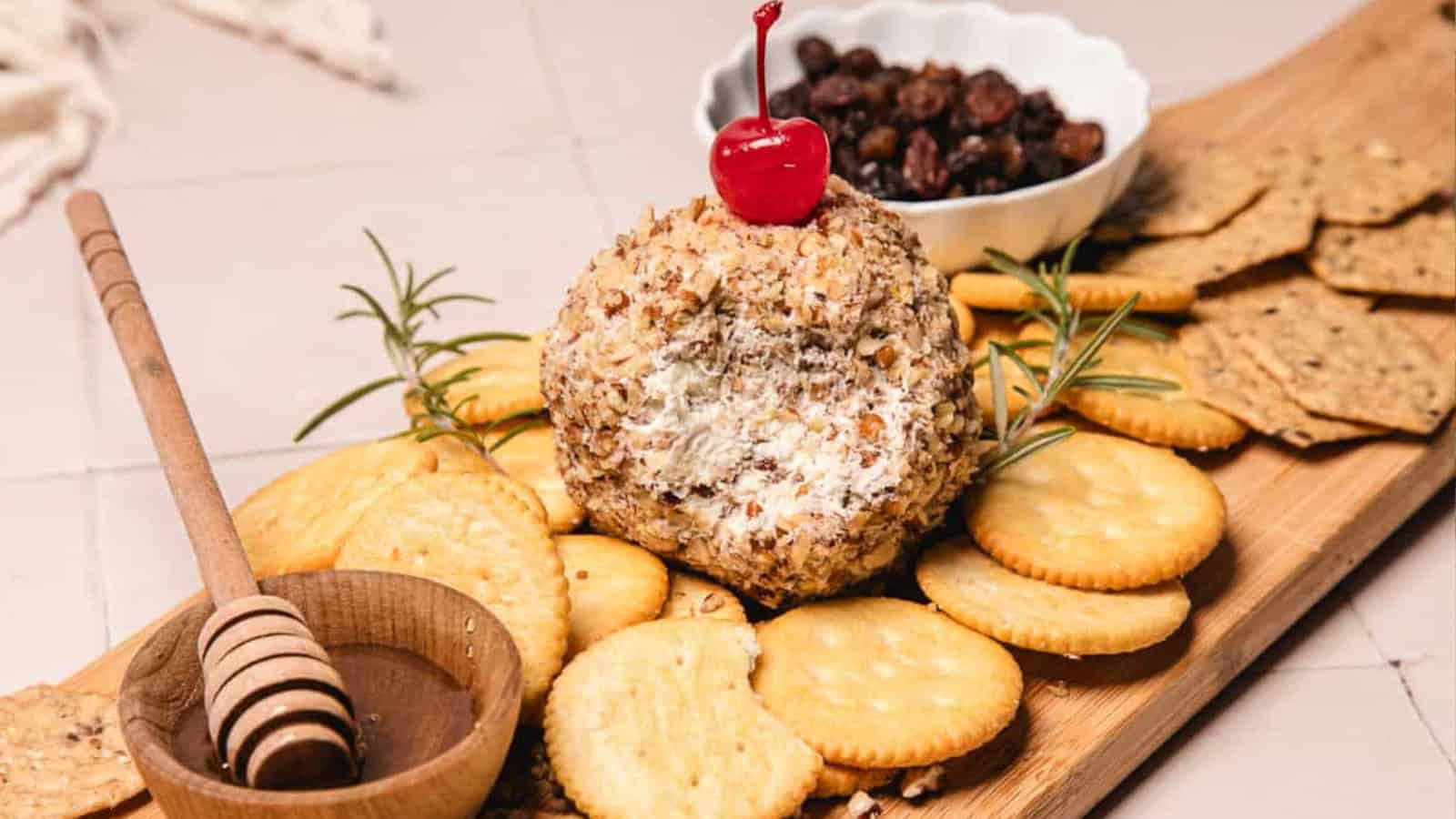 A cheese ball coated with nuts, topped with a cherry, served with crackers, honey, and raisins on a wooden board.