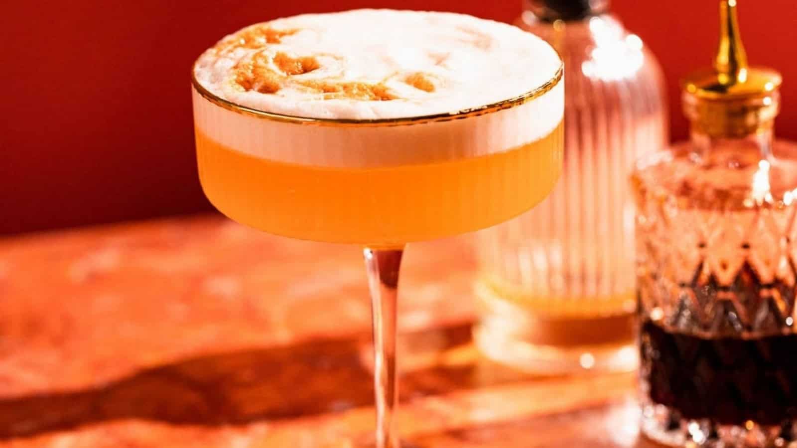 A frothy orange cocktail in a coupe glass on a reddish marble table, with bitters bottles beside it.