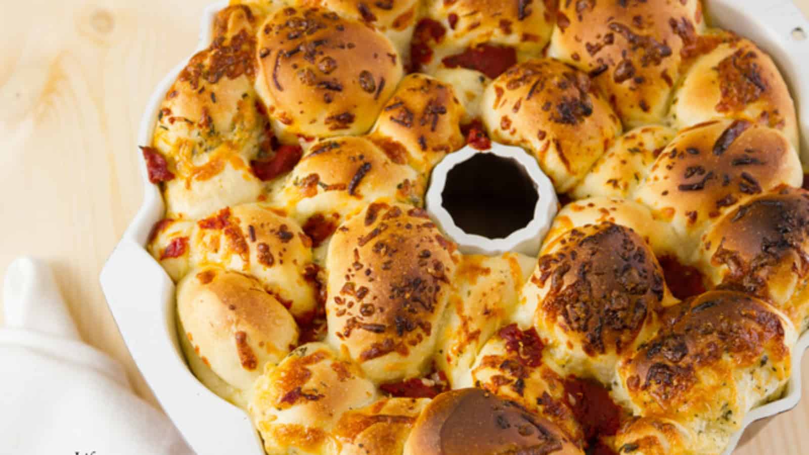 Golden, cheesy pull-apart bread with herbs and pepperoni in a white bundt pan on a light wooden surface.