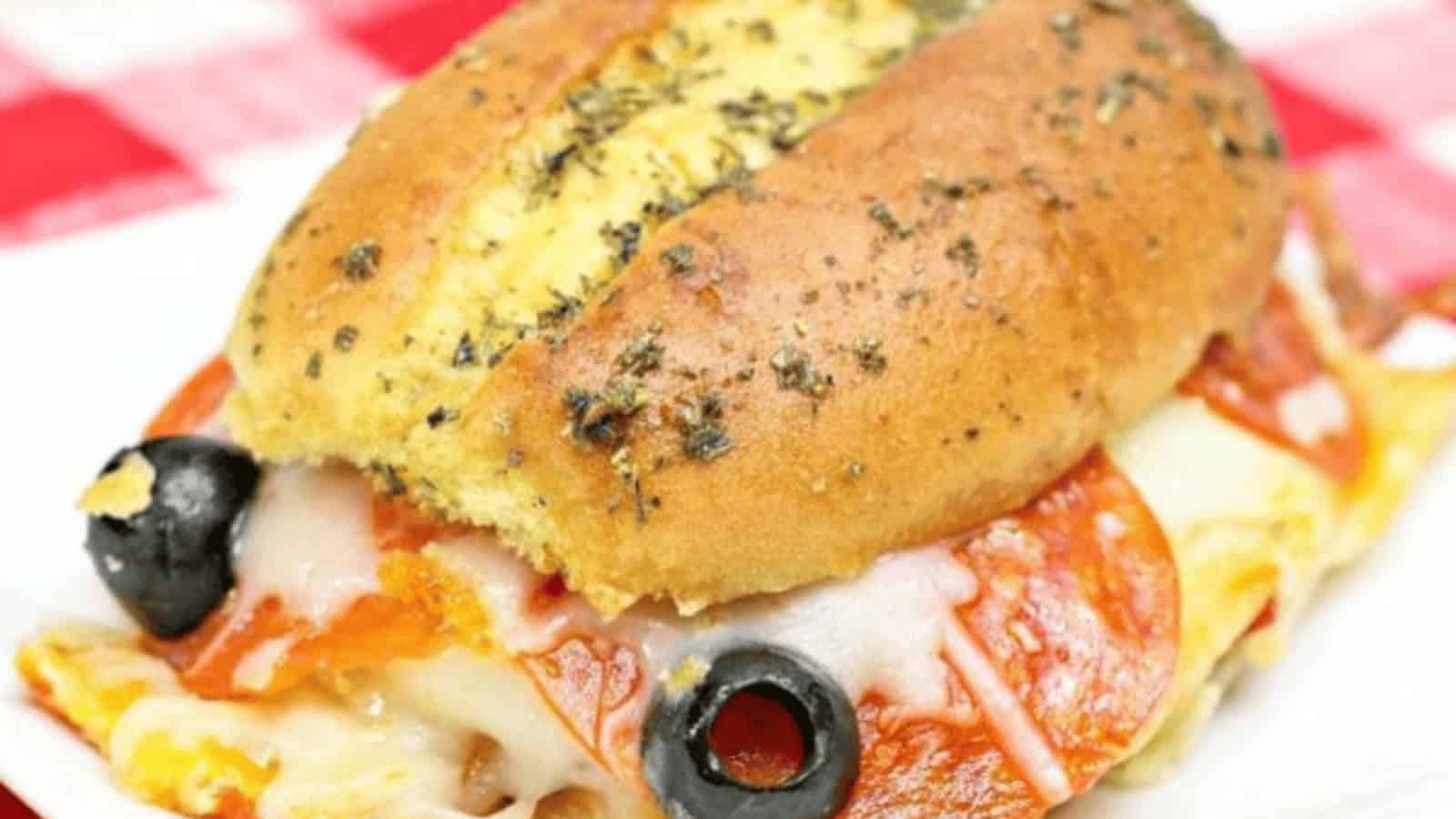 A sandwich with melted cheese, pepperoni, olives, and a seasoned bun on a red-and-white checkered tablecloth.