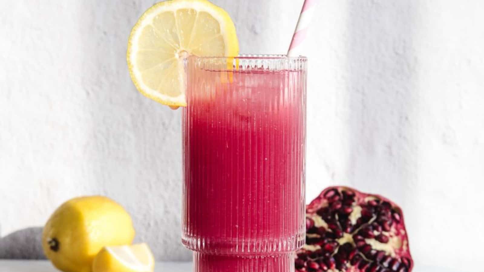 A pink drink with a lemon slice and straw, next to lemons and a halved pomegranate on a white surface.