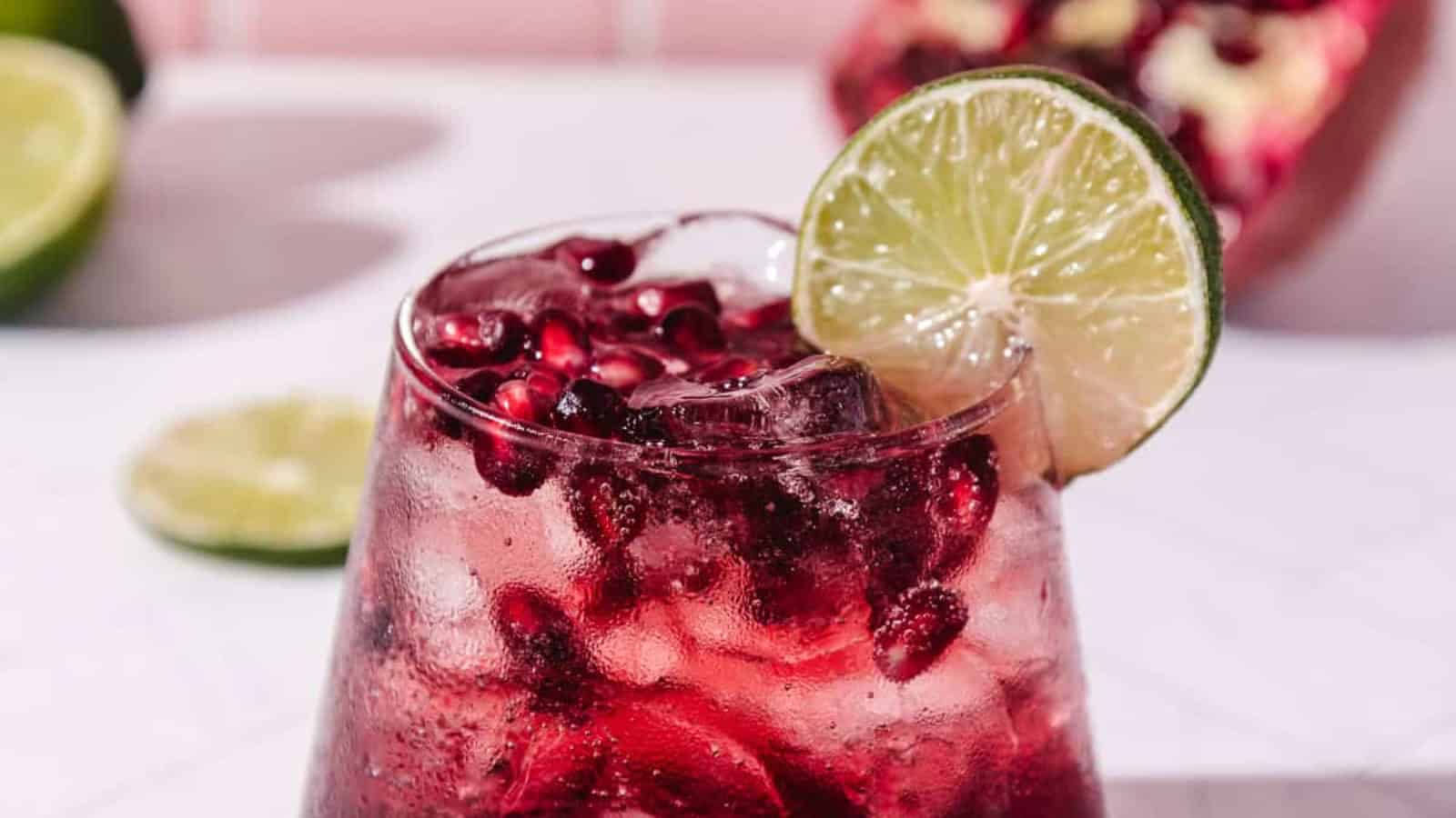 A glass of pomegranate cocktail with ice, pomegranate seeds, and a lime slice garnish.