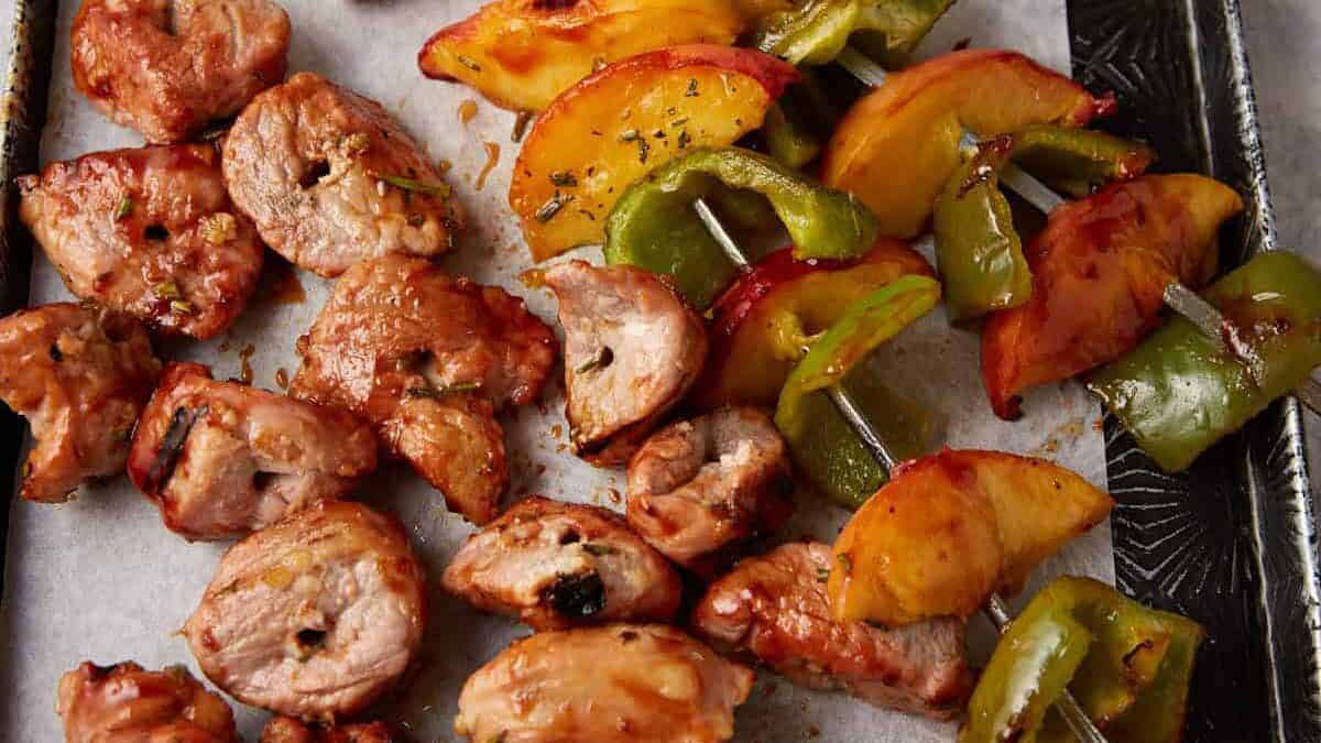 Grilled meat and vegetable skewers with pieces of pork, green bell pepper, and yellow sliced fruit on a parchment-lined tray.
