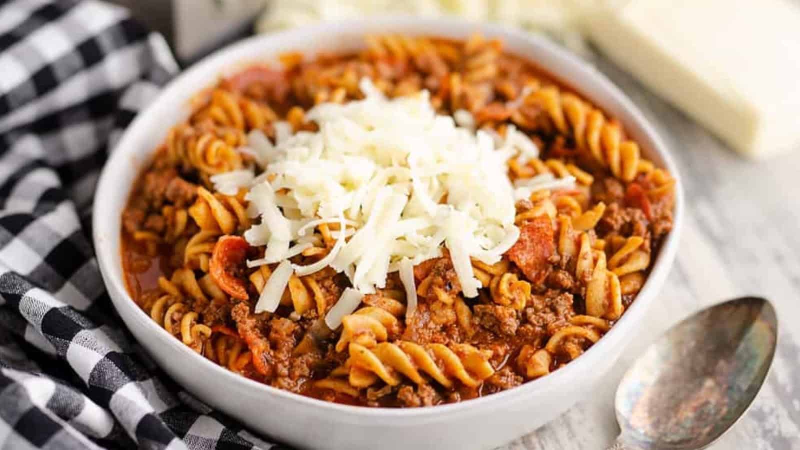 A bowl of rotini pasta with meat sauce, topped with shredded cheese, next to a spoon and checkered napkin.
