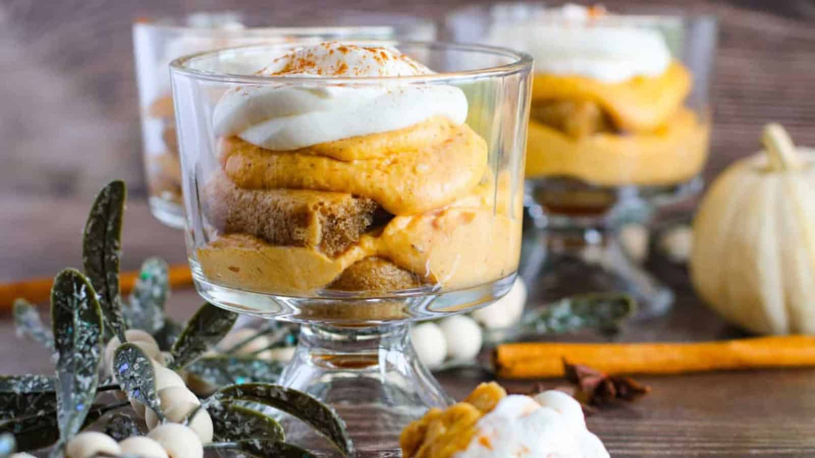 Three glass dessert cups filled with layers of pumpkin mousse, whipped cream, and sponge cake sit on a wooden surface, adorned with greenery and cinnamon sticks. A small white pumpkin is in the background.