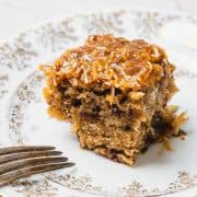 A square piece of German chocolate cake with coconut topping on a decorative plate next to a fork.