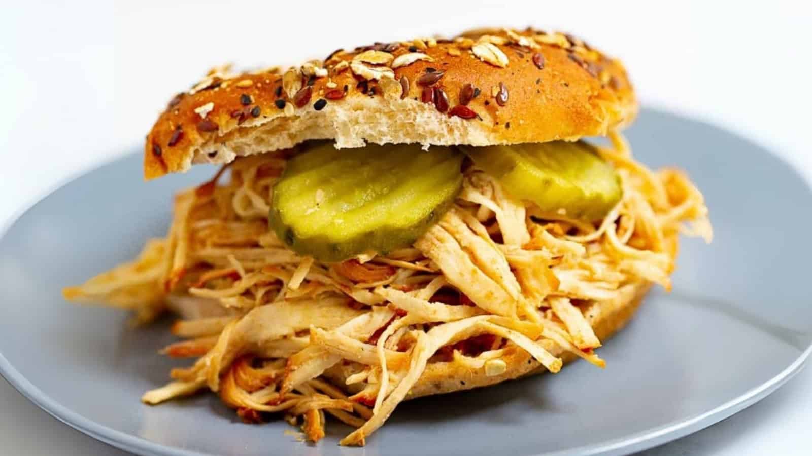 Shredded chicken sandwich with pickles on a seeded bun, served on a gray plate.