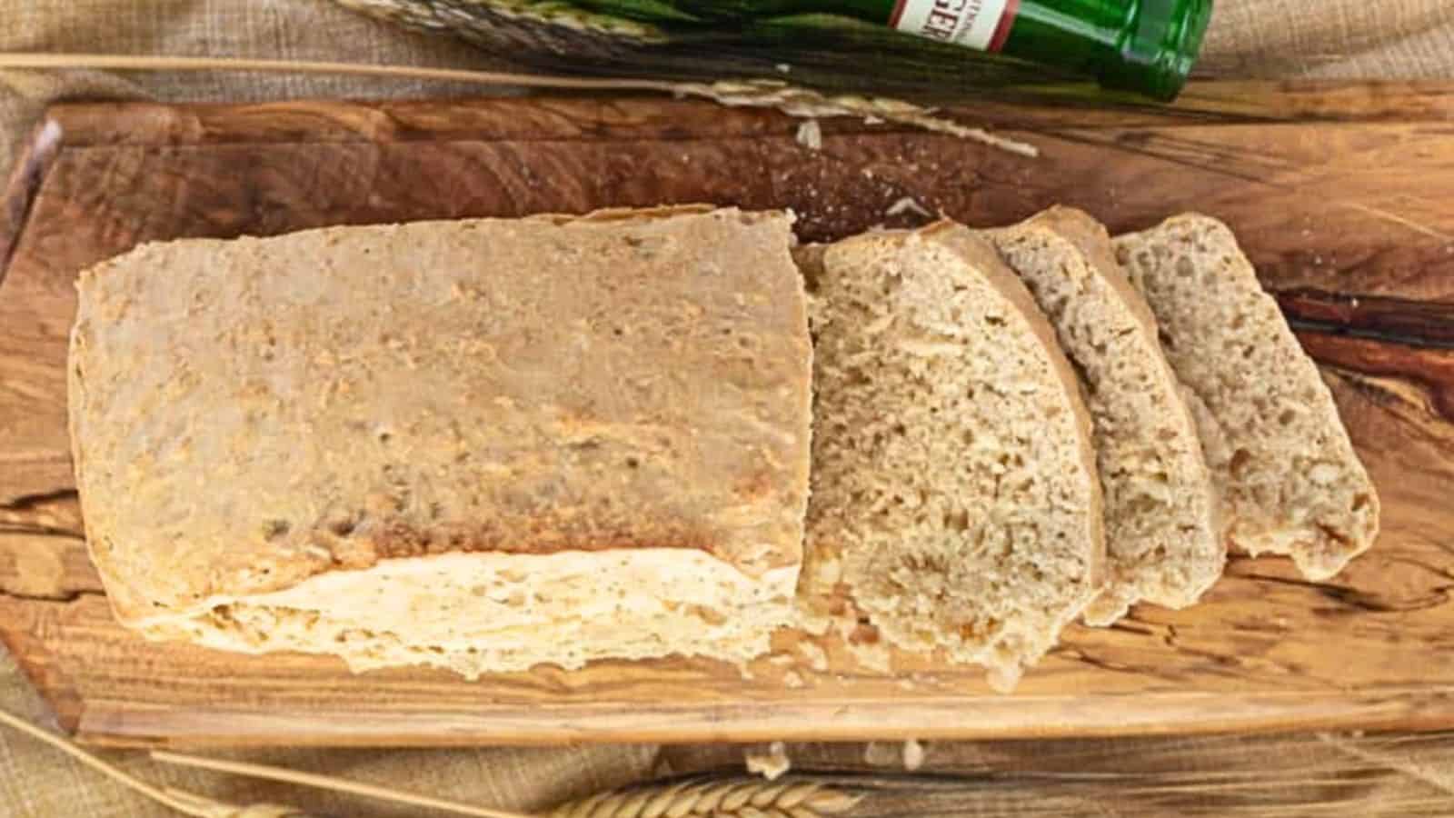 A loaf of homemade bread with three slices cut, displayed on a wooden board next to a green bottle.