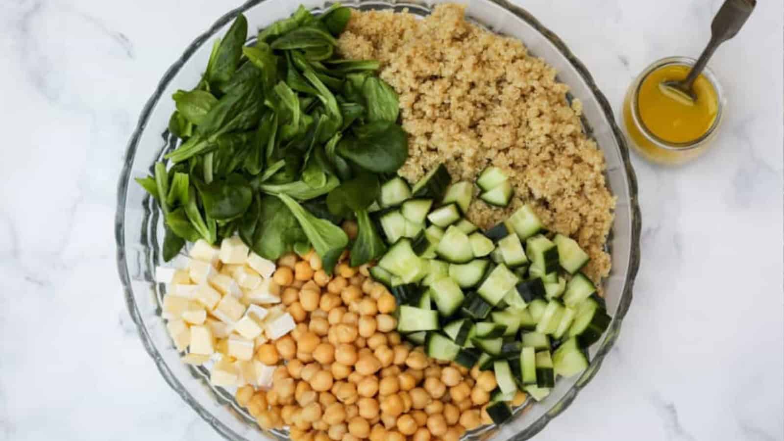 A bowl with spinach, quinoa, chopped cucumber, chickpeas, feta cheese, and a jar of dressing on the side.