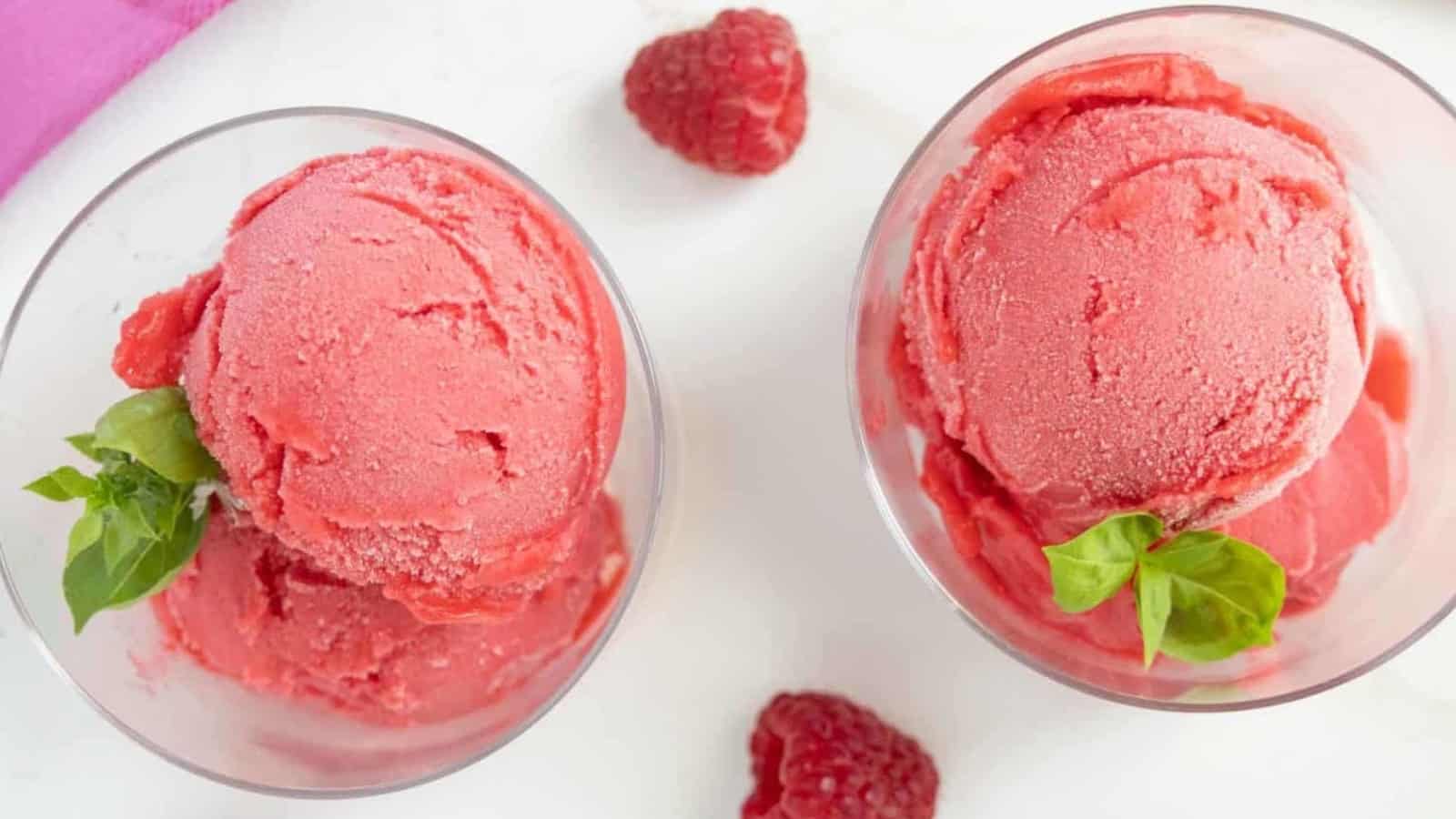 Two glass bowls of raspberry sorbet with fresh raspberries and basil leaves on a white surface.