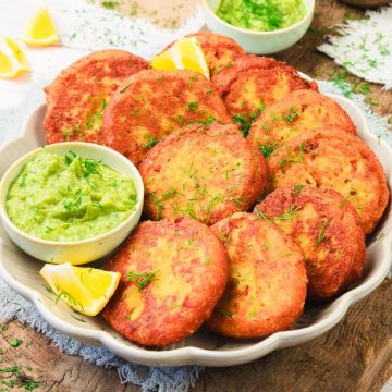 A plate of golden red lentil patties with green dip and lemon wedges, garnished with fresh herbs.