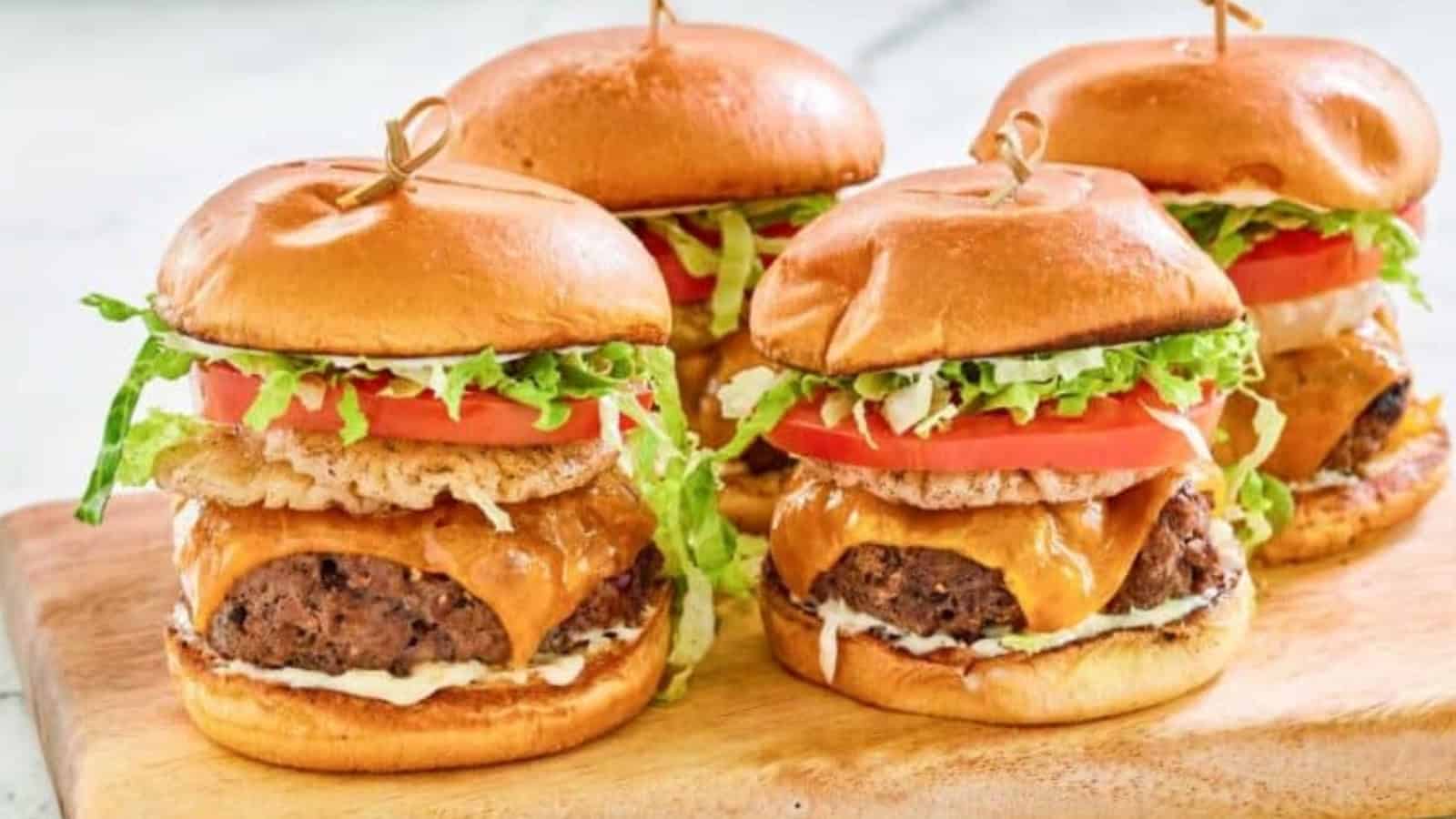 Four cheeseburgers with lettuce, tomato, onion, cheese, and sauce on a wooden board.