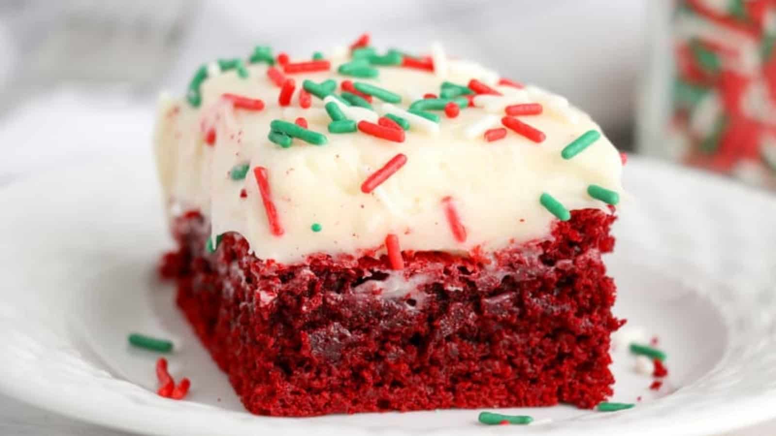 A slice of red velvet cake with white frosting and red and green sprinkles on a white plate.