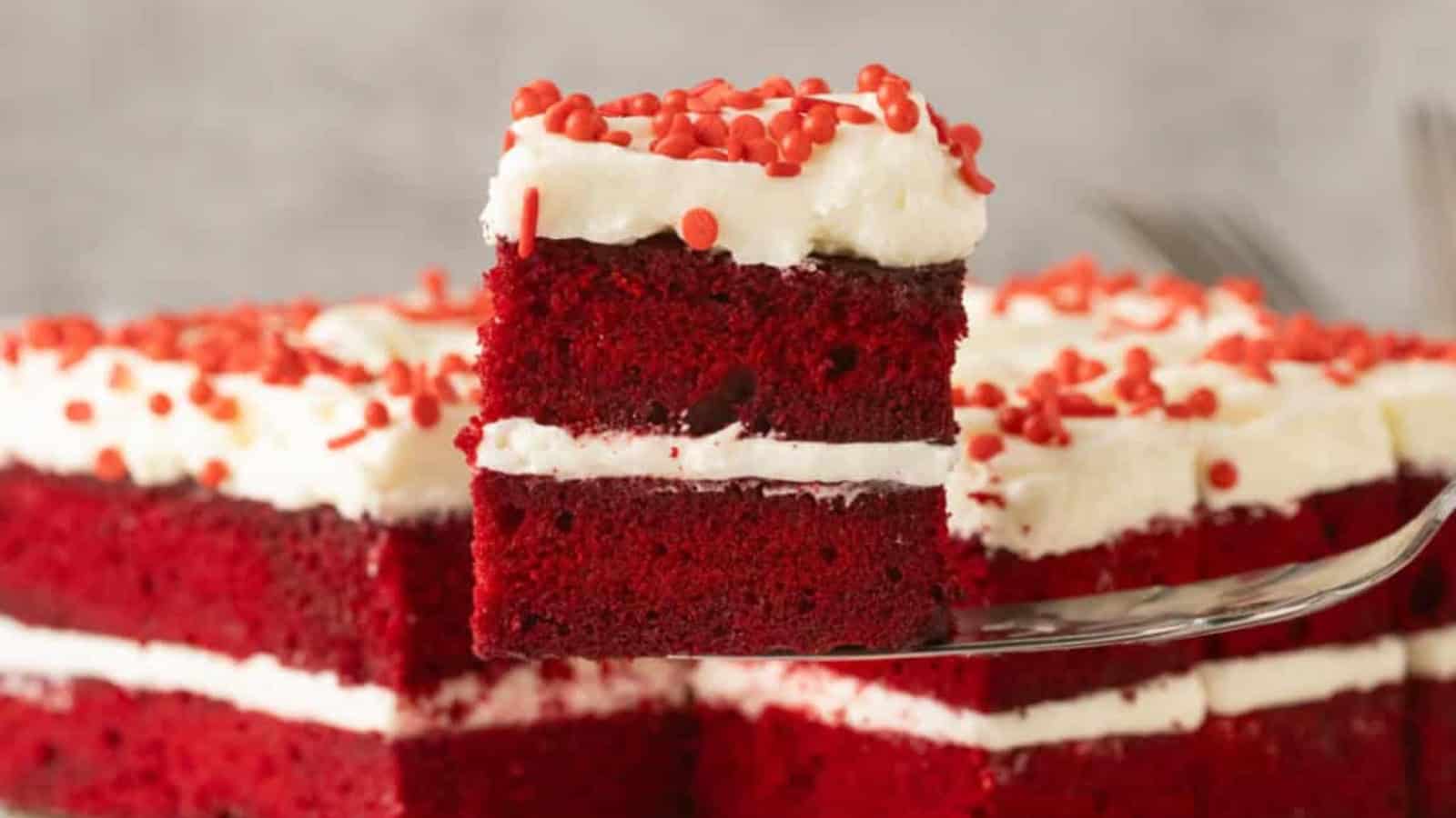 A slice of red velvet cake with white frosting and red sprinkles on a serving spatula.