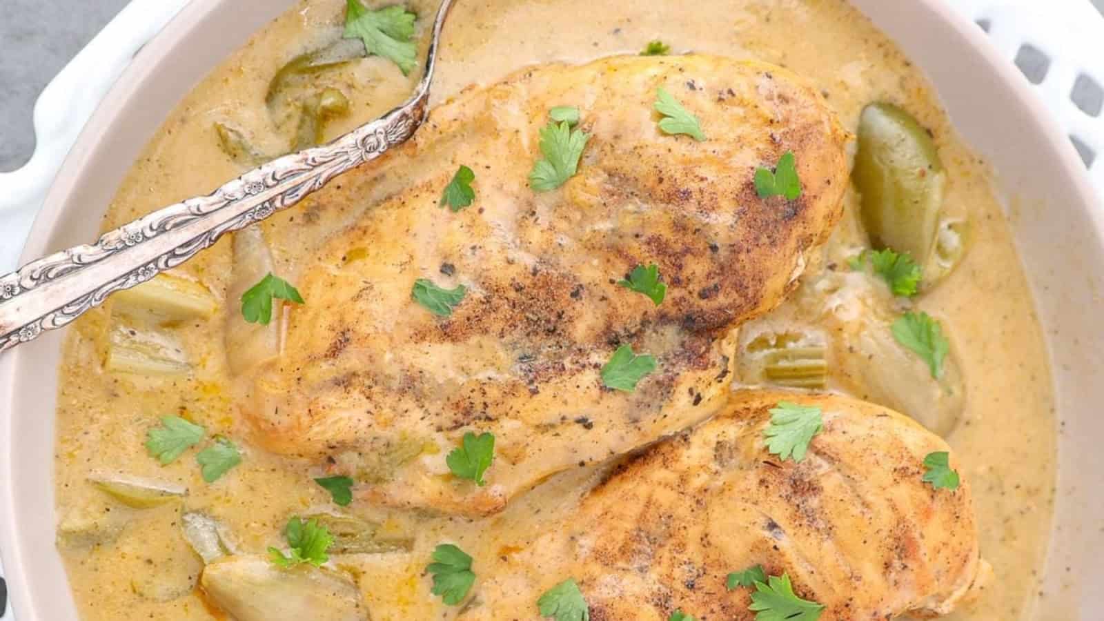 Two cooked chicken breasts in creamy sauce, garnished with cilantro, in a white dish with a spoon.