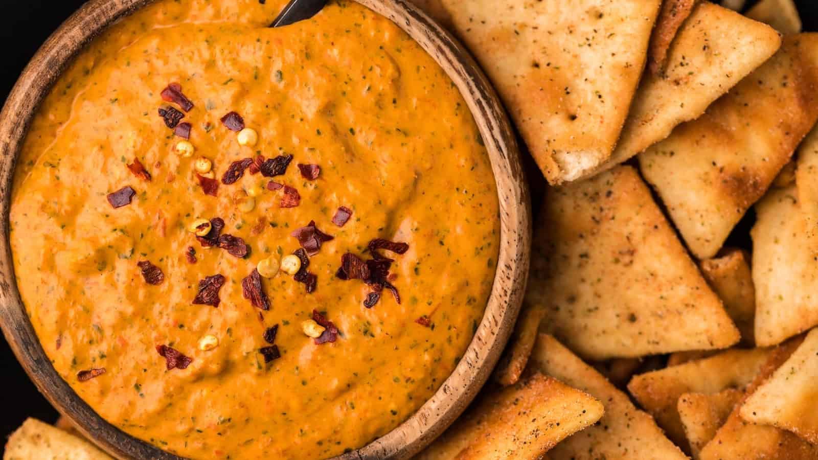 A bowl of creamy red dip topped with chili flakes, served with crispy pita chips.