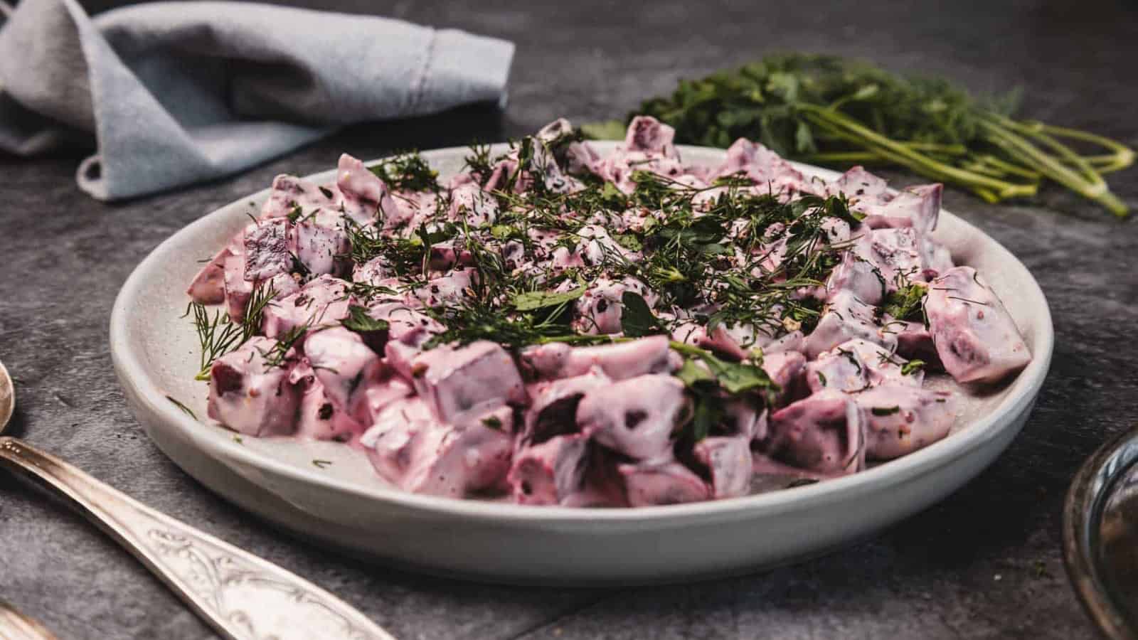 A bowl of creamy beet salad topped with fresh herbs, served on a gray table with utensils and parsley nearby.