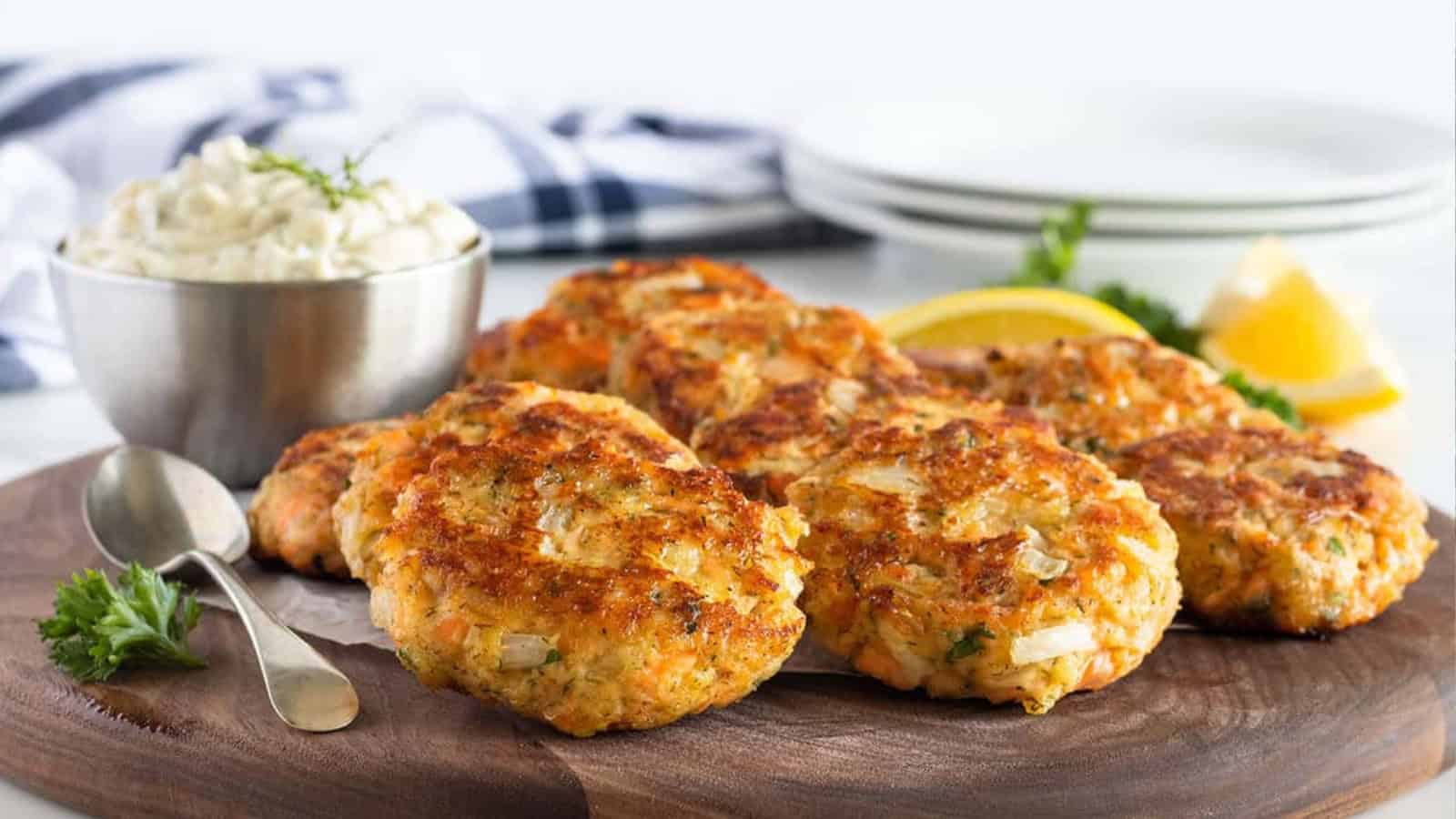 Crab cakes on a wooden board with a bowl of sauce, lemon wedges, and parsley garnish.