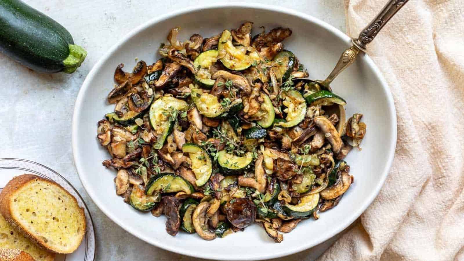 Saut&eacute;ed zucchini and mushrooms garnished with herbs in a white bowl with a spoon.