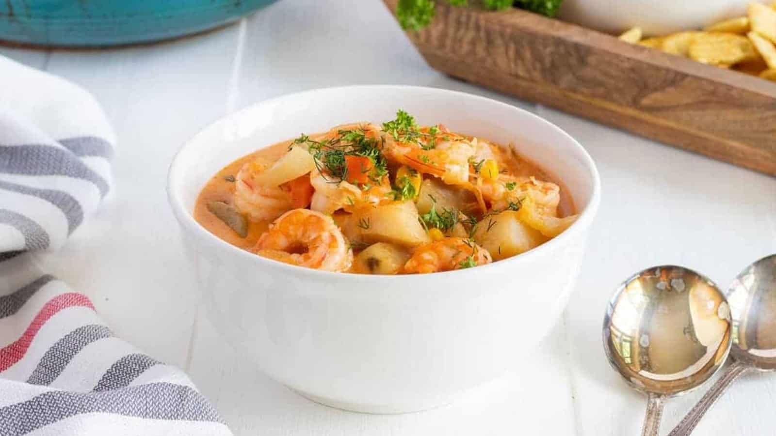 White bowl of creamy seafood stew with shrimp and herbs, placed on a white table beside two spoons.