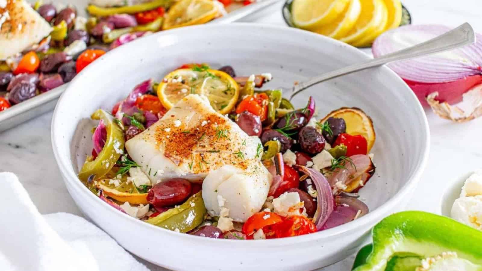 A bowl of baked white fish with olives, tomatoes, onions, peppers, lemon slices, and fresh herbs.