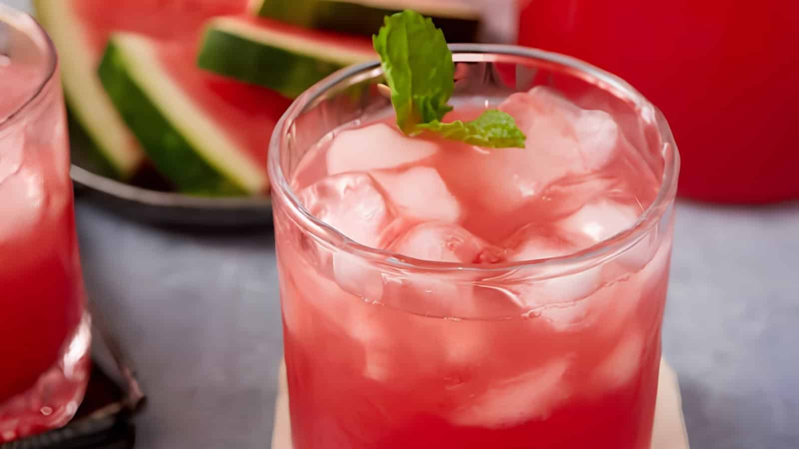 A glass of watermelon drink with ice and mint garnish, with watermelon slices in the background.