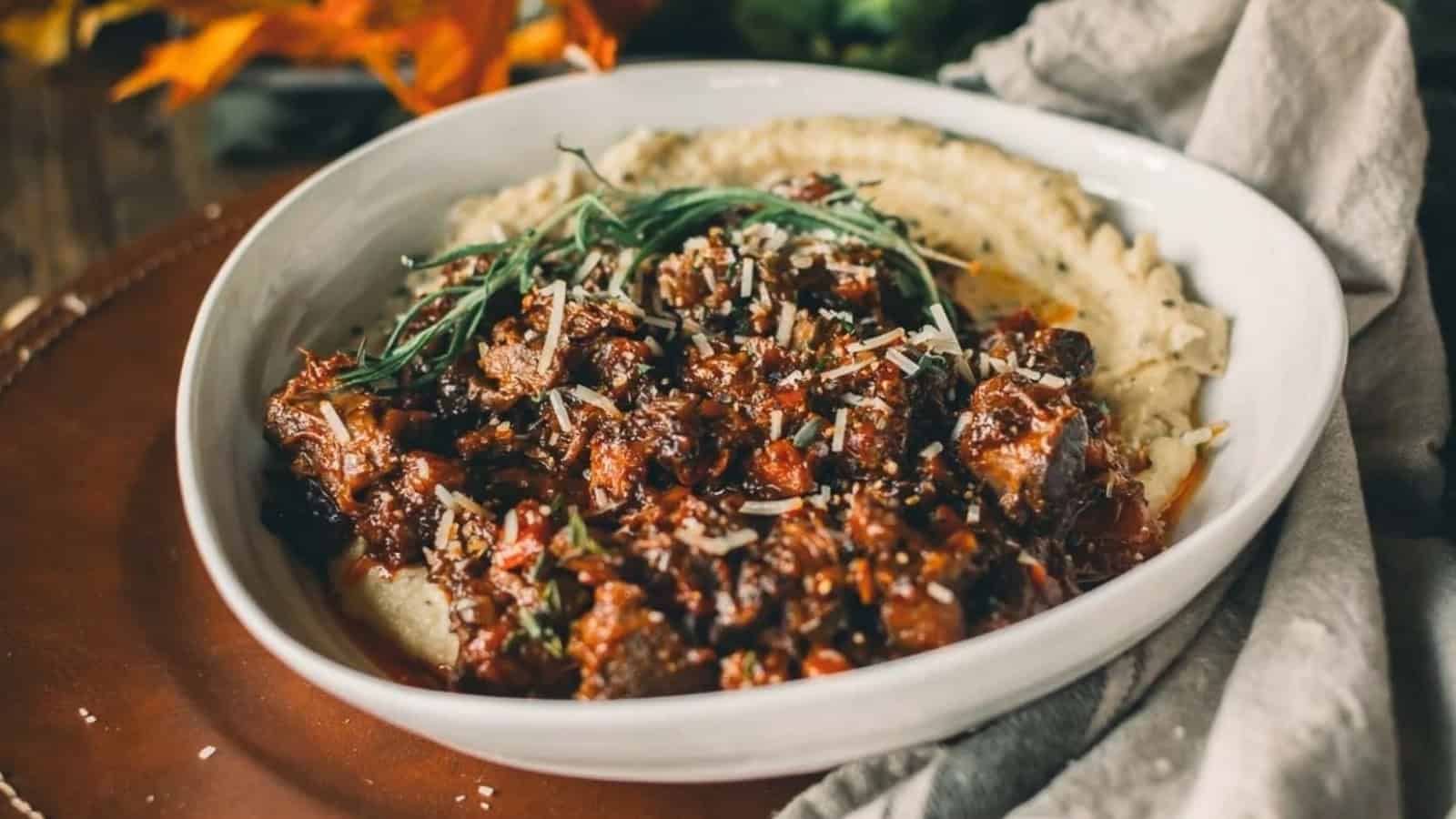 A bowl of braised meat ragu garnished with herbs, served over creamy mashed potatoes.