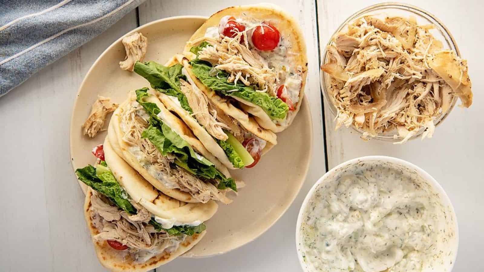 Four pita wraps with shredded chicken, lettuce, tomatoes, and sauce on a plate, with extra chicken and sauce on the side.
