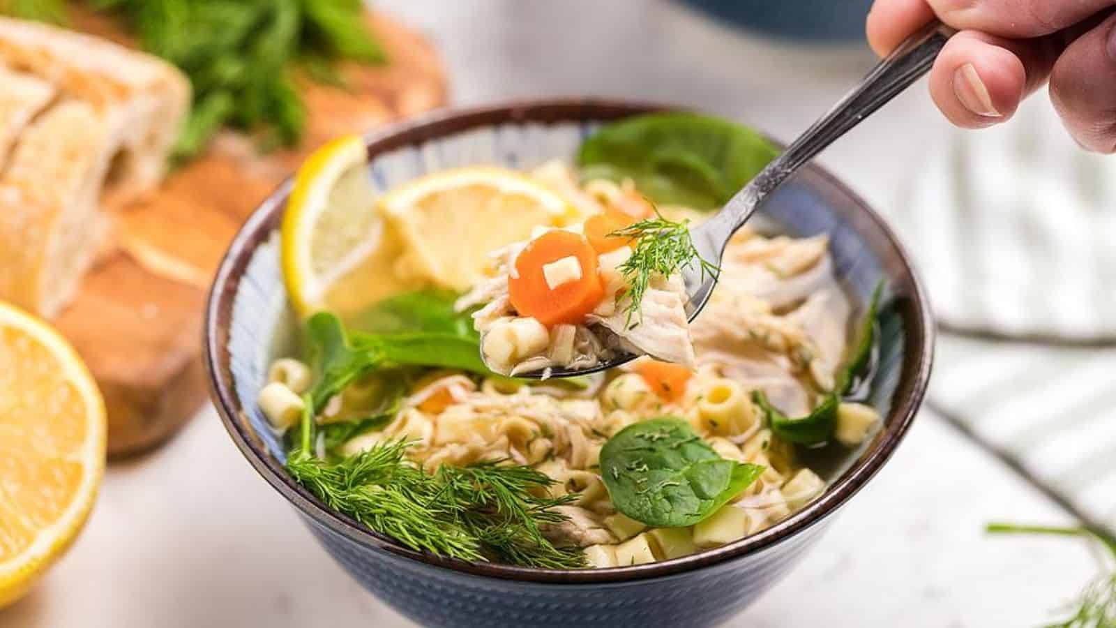 Bowl of chicken noodle soup with carrots, spinach, dill, and lemon slices; a spoonful is being lifted.