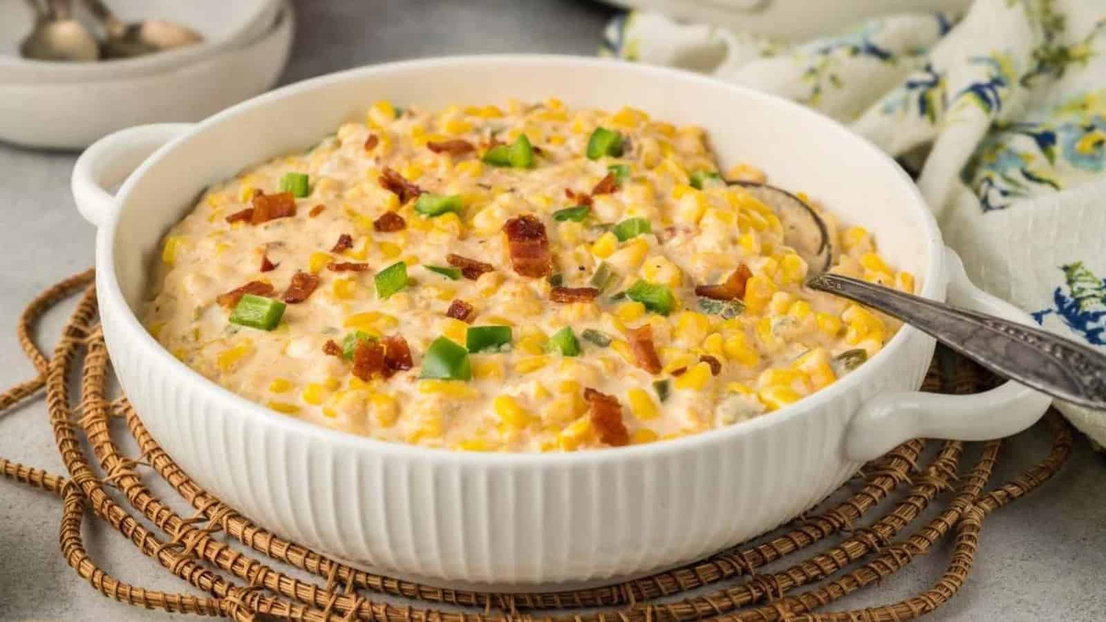 Creamy corn casserole topped with green peppers and bacon bits in a white baking dish with a spoon.