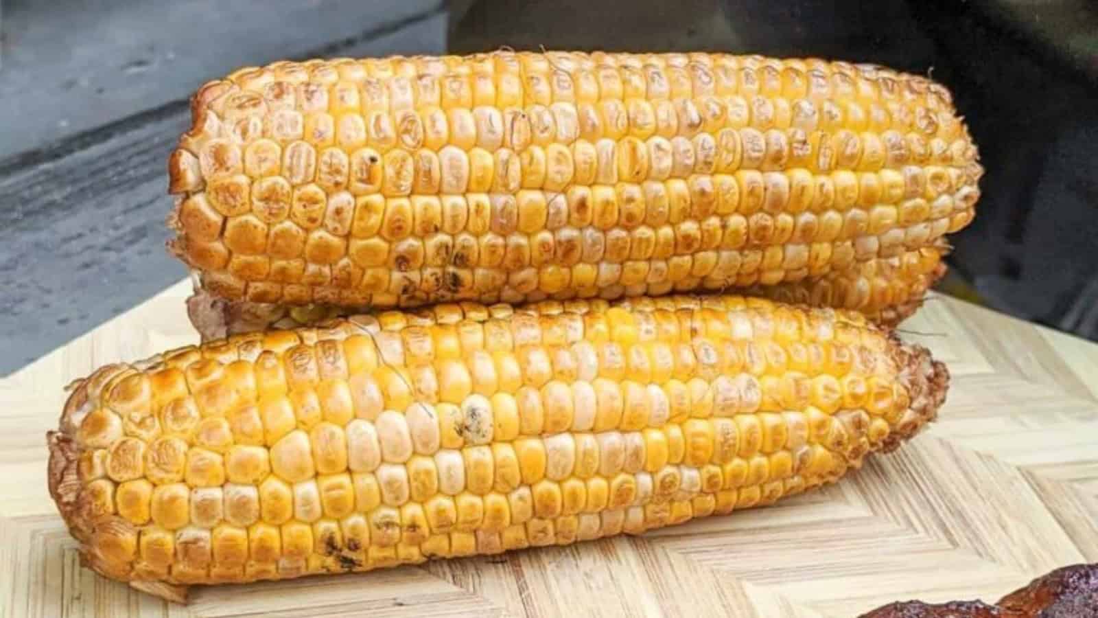 Two grilled corn cobs stacked on a wooden board, showing golden, slightly charred kernels.