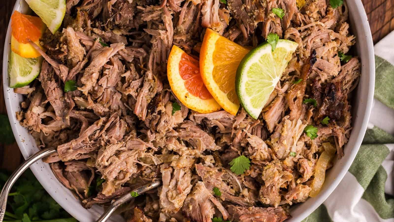 Shredded pork carnitas garnished with orange and lime slices in a bowl, with metal serving tongs.