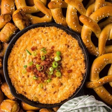 A skillet of cheesy dip topped with bacon and green onions, surrounded by pretzels and pretzel bites.