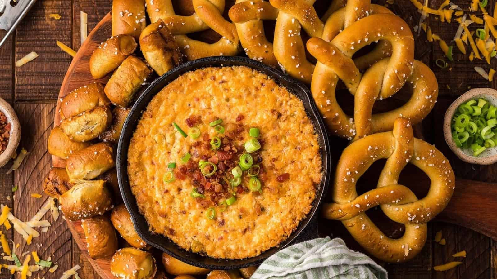 A skillet of cheesy dip topped with bacon and green onions, surrounded by pretzels and pretzel bites.